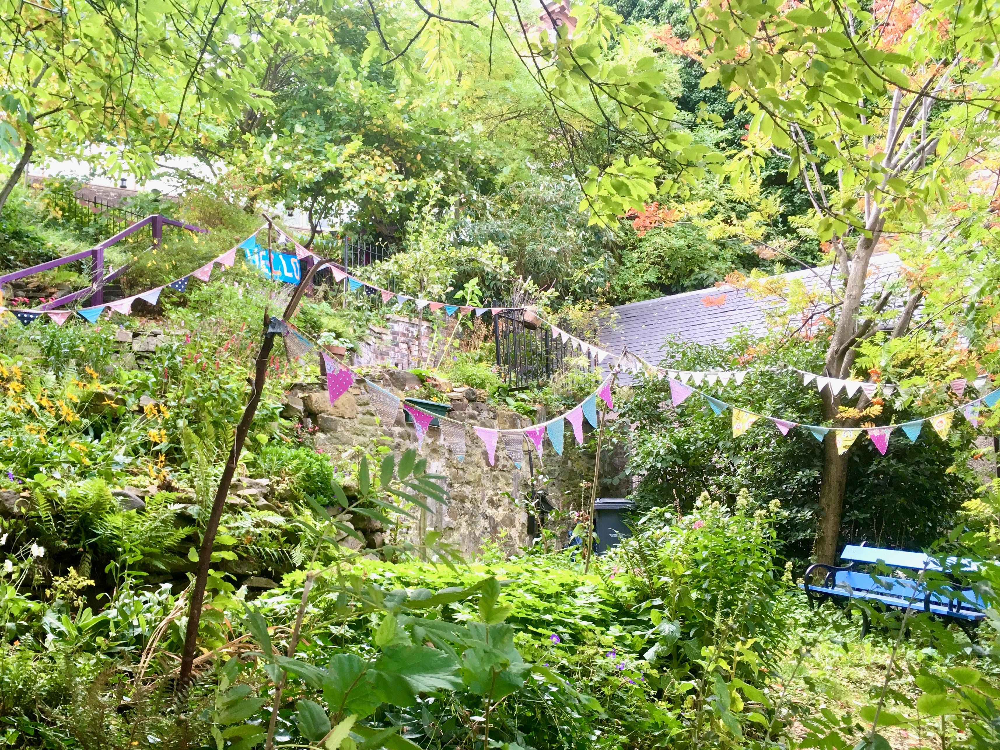 Green garden with stone wall and colorful flags, surrounded by trees and foliage.